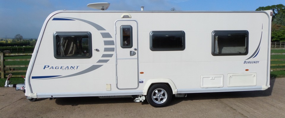 A Bailey Pageant Burgundy touring caravan in the yard, with fields and trees behind